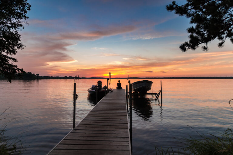 Beautiful lake sunrise sunset with a young woman sitting on the end of a dock, with a boats. on either side. Colorful tranquil vacation scene with concepts of peacefulness and beauty